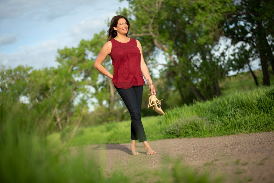 Tiffany Edgar walking barefoot on a Colorado trail, shoes in hand, smiling